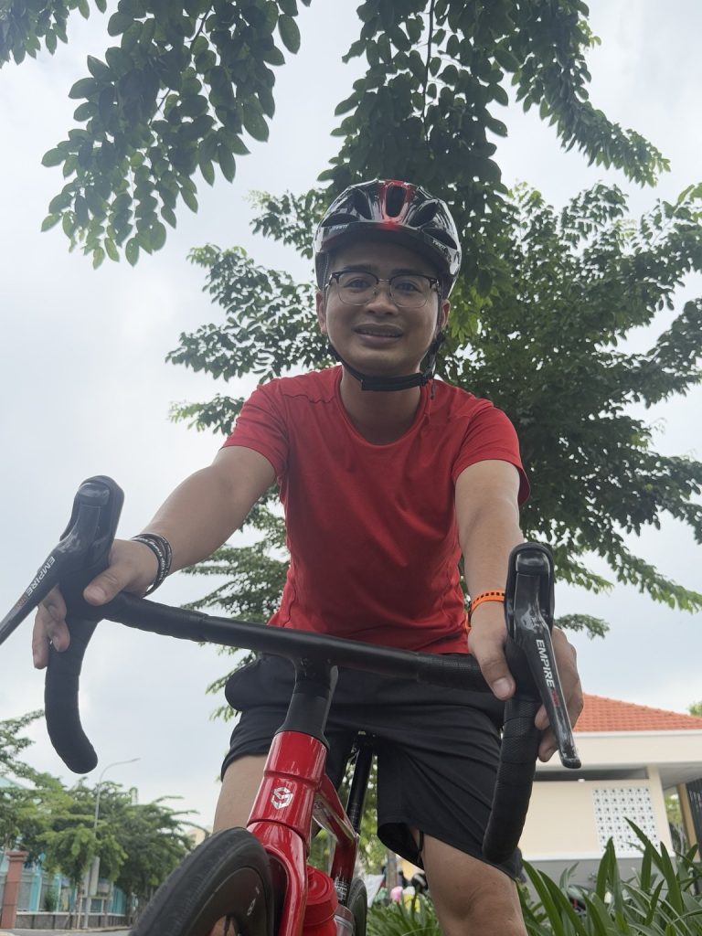 Lê Tấn Đào đội mũ bảo hiểm, mặc áo đỏ cùng chiếc roadbike đỏ dưới tán cây