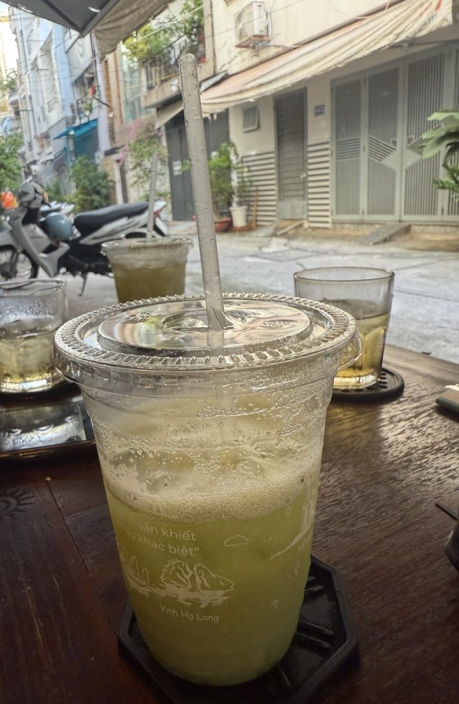 Ly nước ép ổi có nắp và ống hút trên bàn gỗ của quán, phía sau là mấy ly nước khác và con hẻm