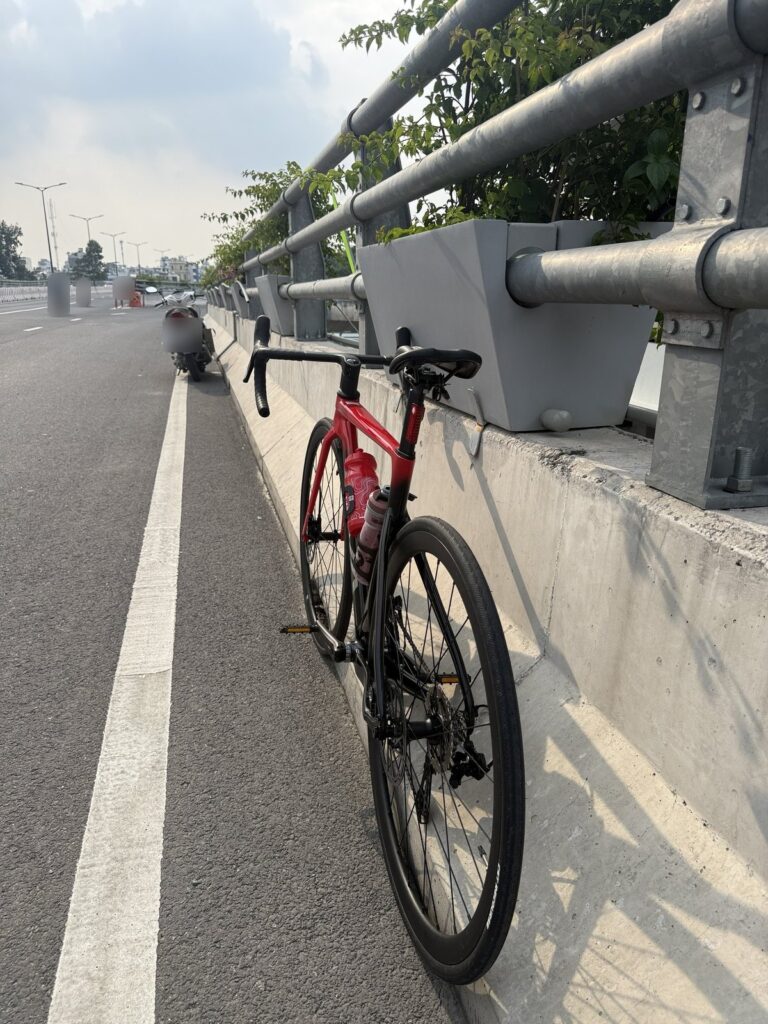 Chiếc xe đạp đường trường màu đỏ đen dựa vào lan can một đoạn đường trên cao, phía sau có xe máy đậu, biển số đã được làm mờ